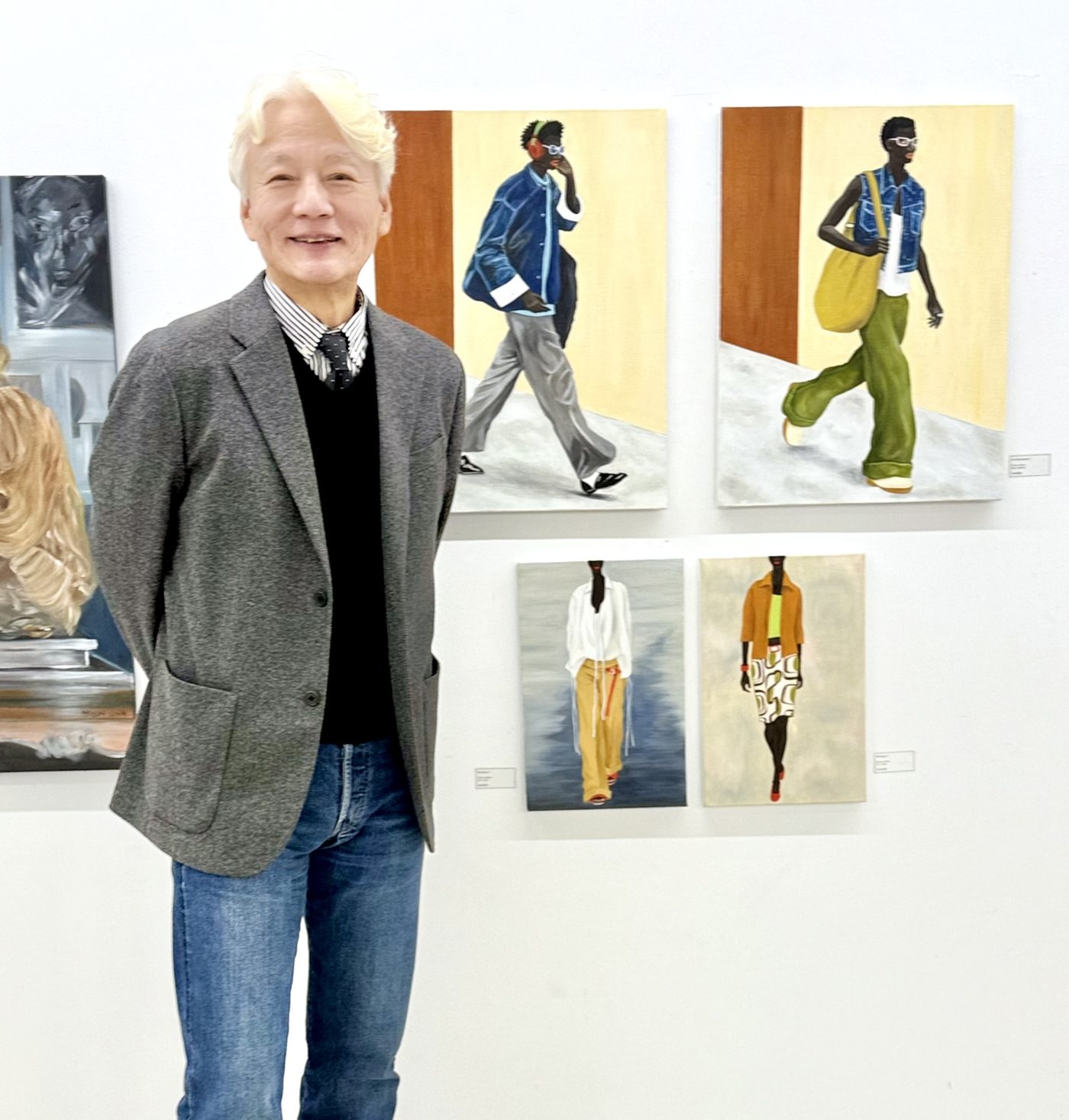 흰 벽에 세로로 긴 인물화 네 점이 두 줄로 걸려 있고, 회색 재킷에 청바지를 입은 백발의 작가가 그 앞에 서서 정면을 보고 있다. 그림 속 인물들은 파란 재킷과 초록 바지, 흰 셔츠와 노란 바지 차림으로 걷고 있으며 작품 옆에는 작은 라벨이 붙어 있다