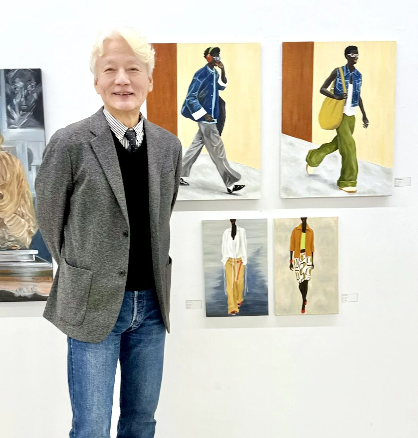 흰 벽에 세로로 긴 인물화 네 점이 두 줄로 걸려 있고, 회색 재킷에 청바지를 입은 백발의 작가가 그 앞에 서서 정면을 보고 있다. 그림 속 인물들은 파란 재킷과 초록 바지, 흰 셔츠와 노란 바지 차림으로 걷고 있으며 작품 옆에는 작은 라벨이 붙어 있다