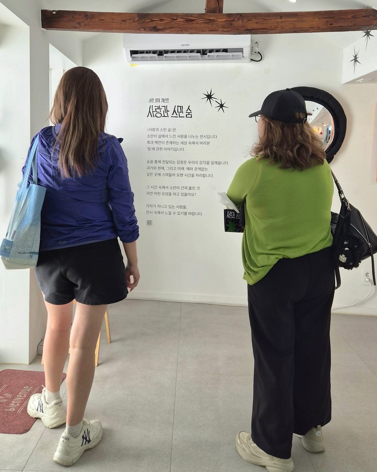 전시장 안쪽 흰 벽에 붙은 전시 서문 앞에서 관람객 두 사람이 뒷모습으로 서서 글을 읽고 있다. 벽에는 전시 제목과 소개 글이 검은 글씨로 인쇄되어 있고 오른쪽에는 둥근 형태의 검은 오브제가 걸려 있다